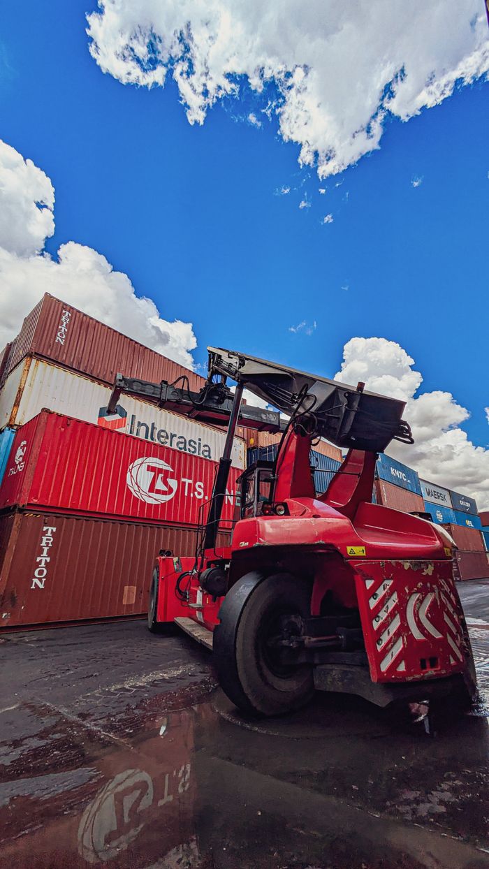 Home Dynamic view of a red forklift amidst stacked shipping containers under a vibrant blue sky.
